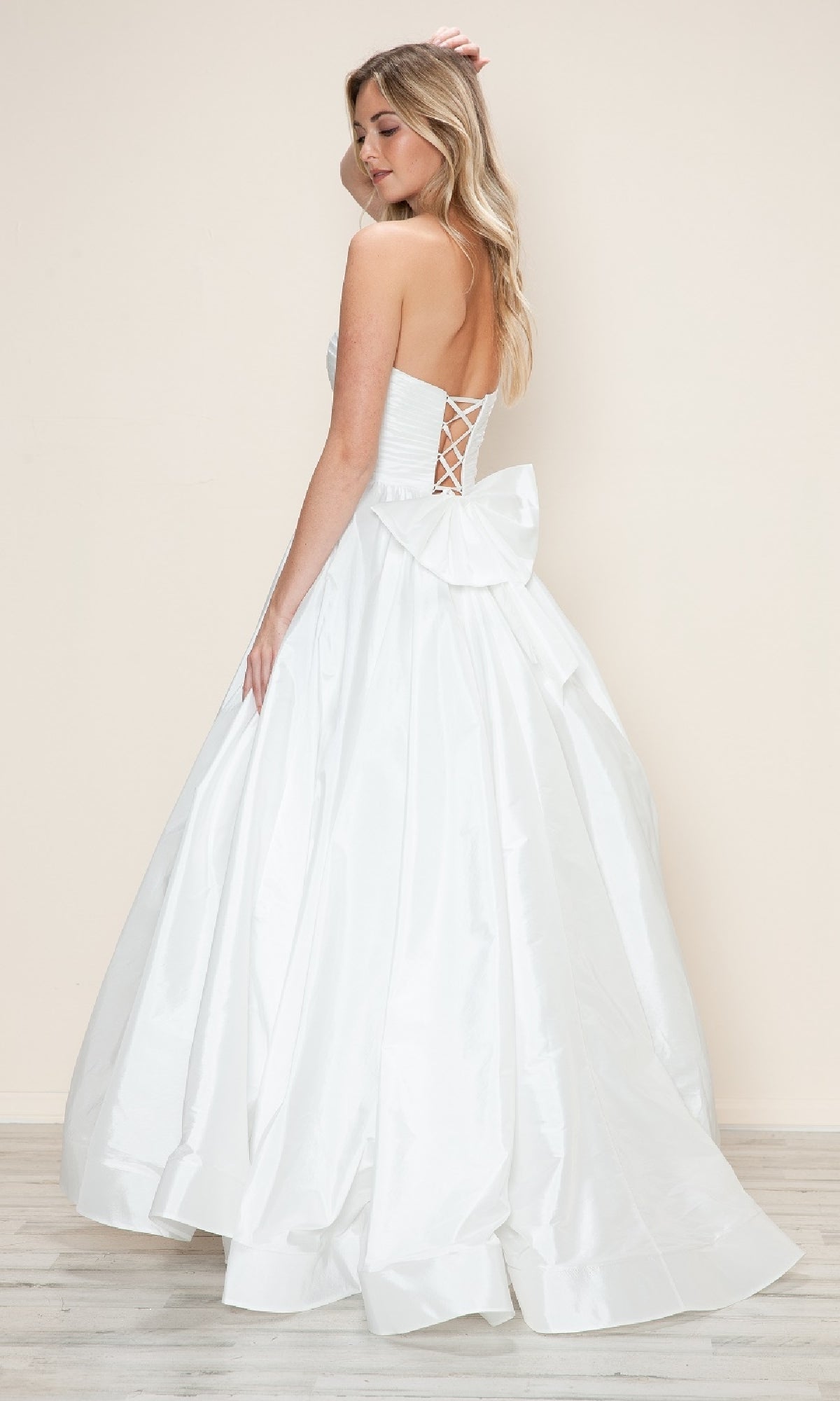 A woman wearing the Long Prom Dress: Poly USA 9754, featuring a strapless ruched bodice, adjustable lace-up back, and large bow, stands on a light wood floor with a plain beige background.